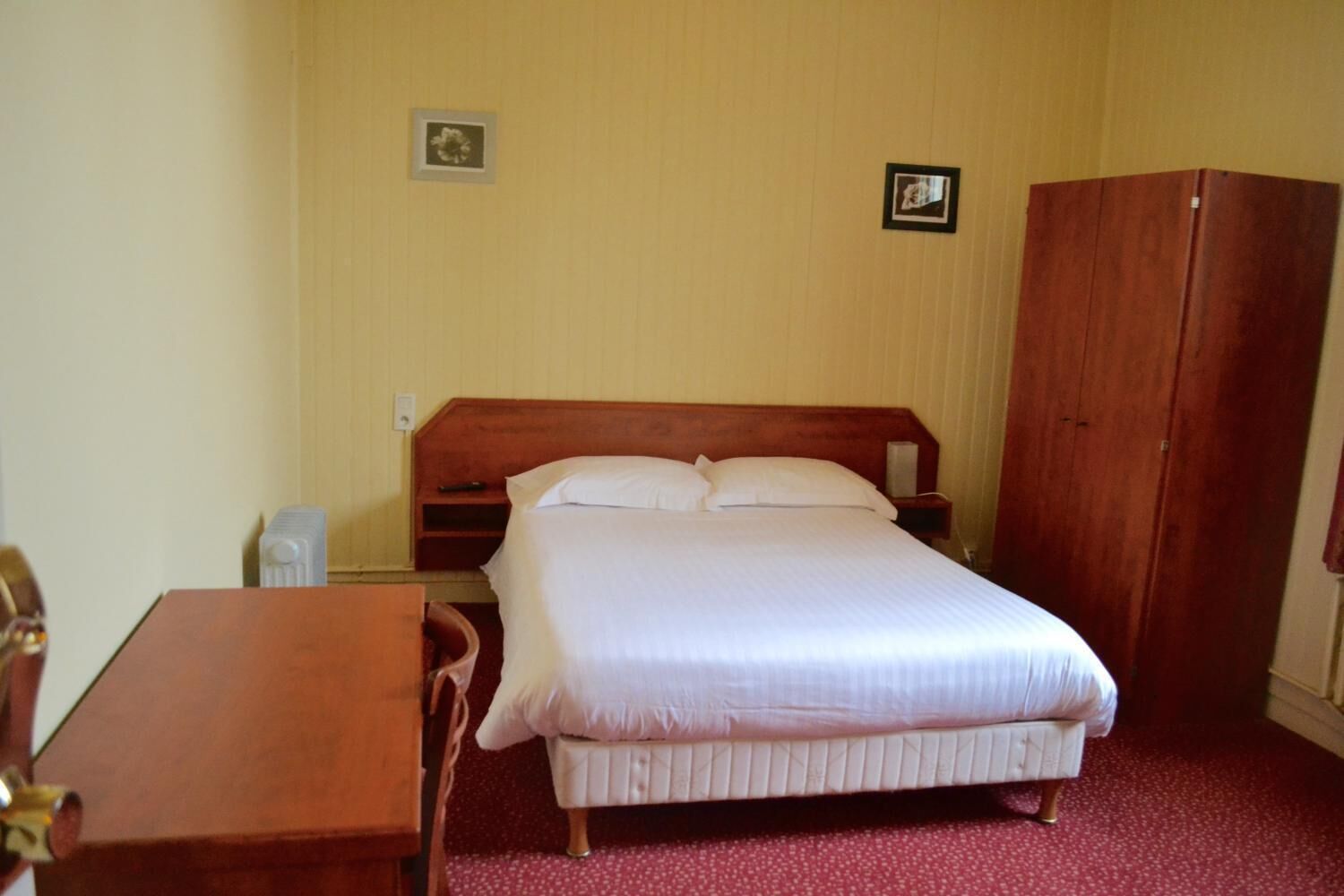 A single bed with a wooden headboard, a bedside table, a chair, and a wardrobe in a room with plain walls.