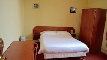 A single bed with a wooden headboard, a bedside table, a chair, and a wardrobe in a room with plain walls.