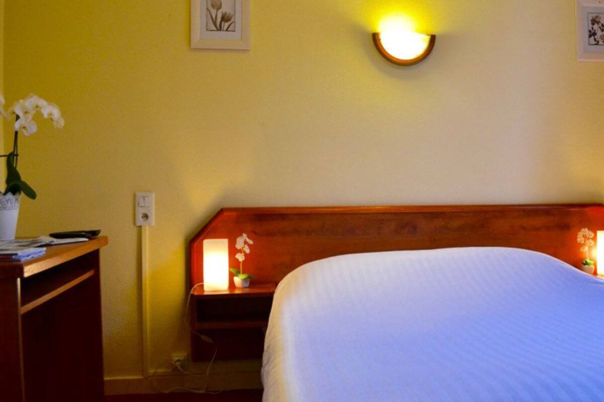 Hotellihuone, jossa on puinen sängynpääty, valkoisilla lakanoiden koristeltu sänky, yöpöytä lampun kanssa ja työpöytä, jossa on kasvi.