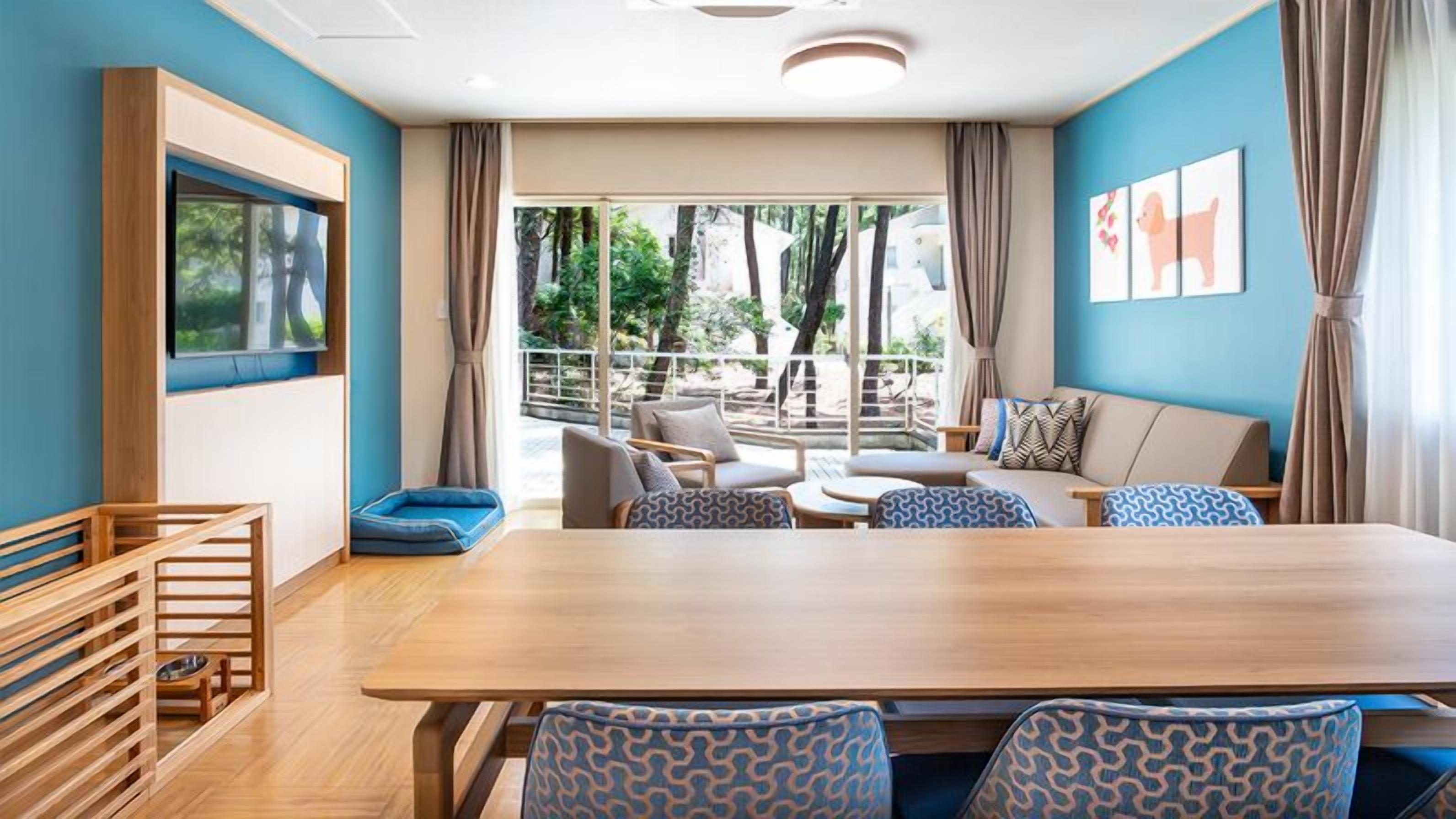 Une chambre d’hôtel moderne dotée d’une grande table à manger en bois, de sièges confortables, d’un couchage pour animaux de compagnie et de grandes fenêtres offrant une vue sur l’extérieur.