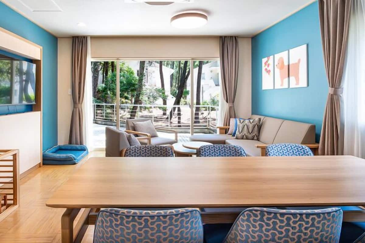 A modern hotel room with a large wooden dining table, comfortable seating, a pet bed, and large windows offering a view of the outdoors.