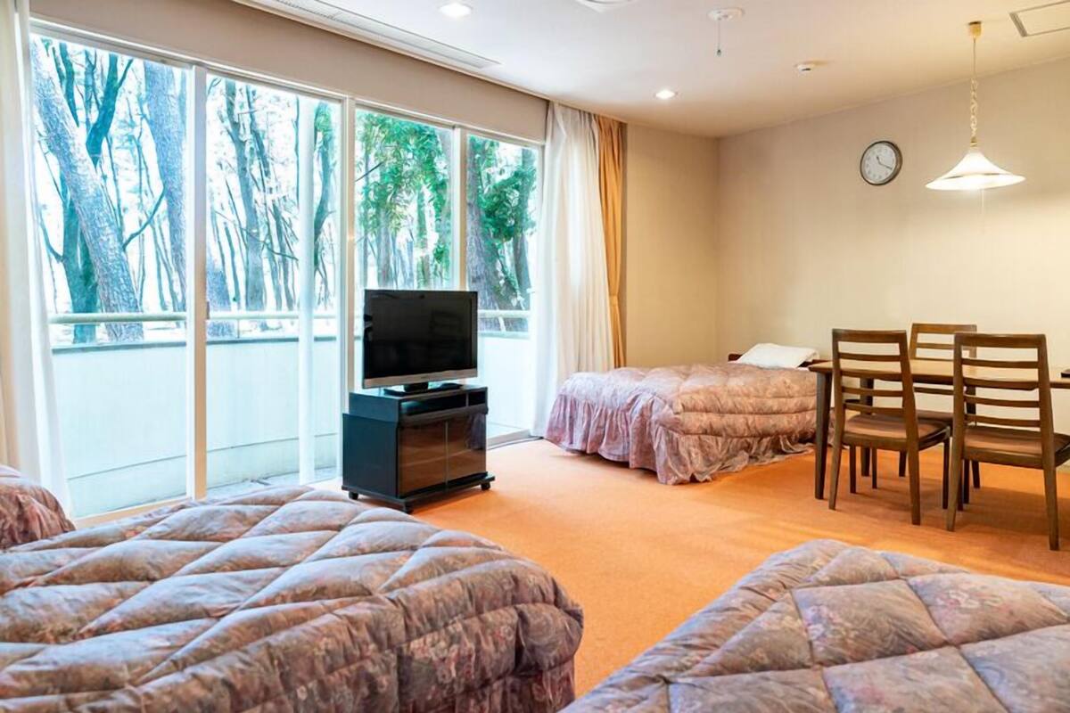 A hotel room with two beds, a TV, a dining table with chairs, and a large window with a view of trees.
