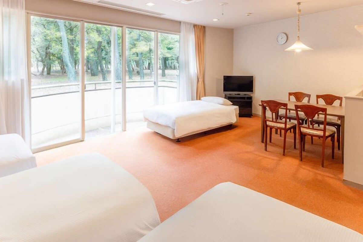 A hotel room with two beds, a dining area with chairs, a TV, and large windows with a view of trees.