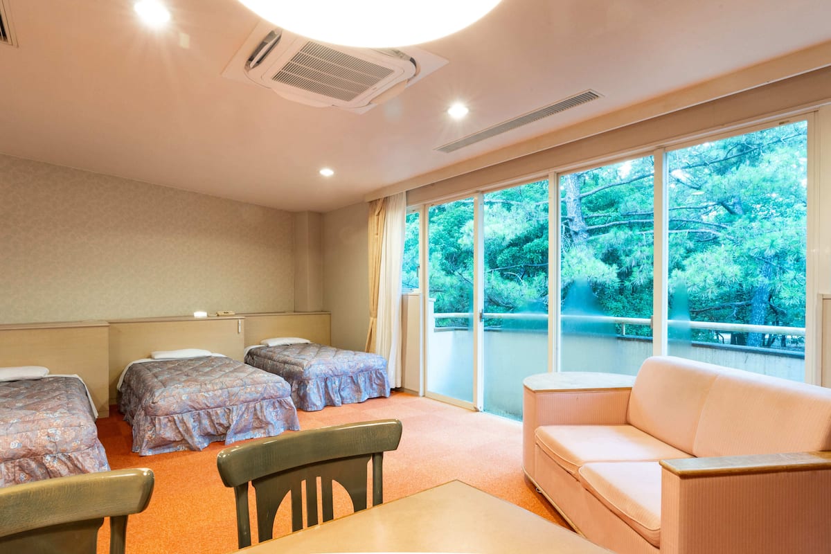 A hotel room with two beds, a sofa, a table, and a large window with a view of trees.