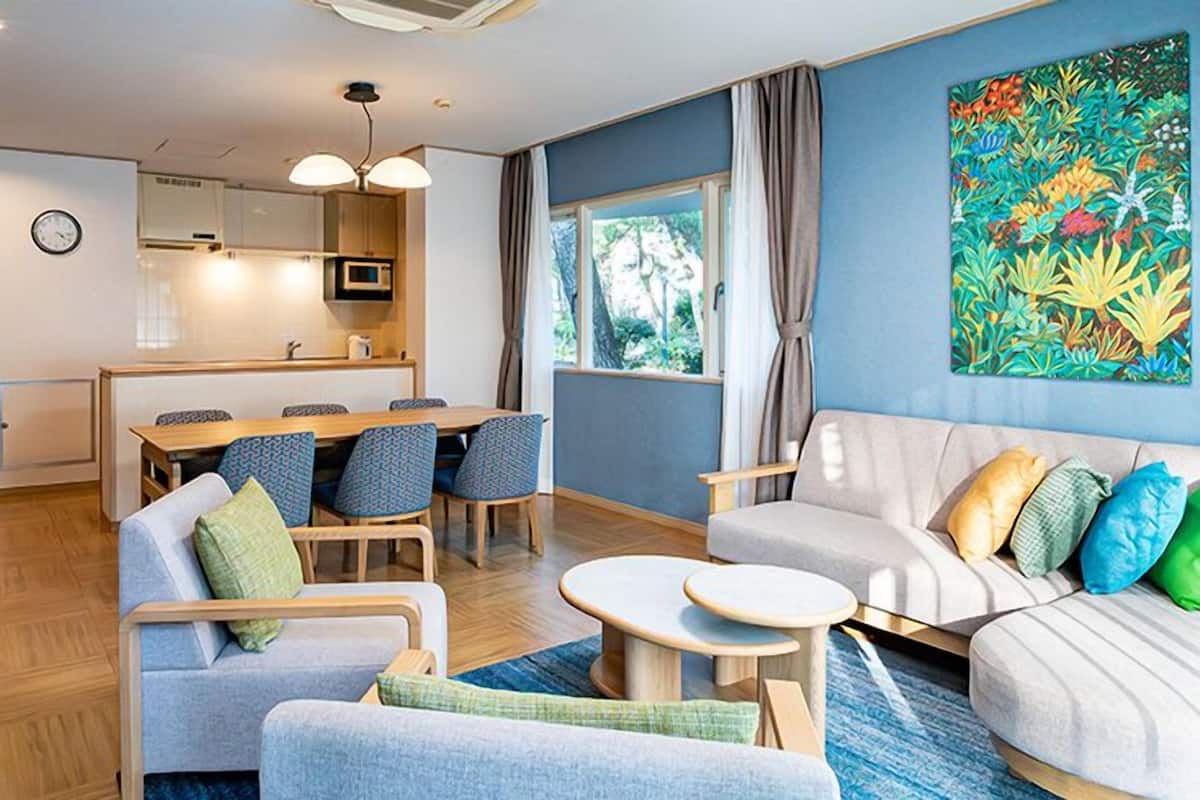 A modern living room with a sofa, a dining table with chairs, a kitchen area, and a large window with curtains.