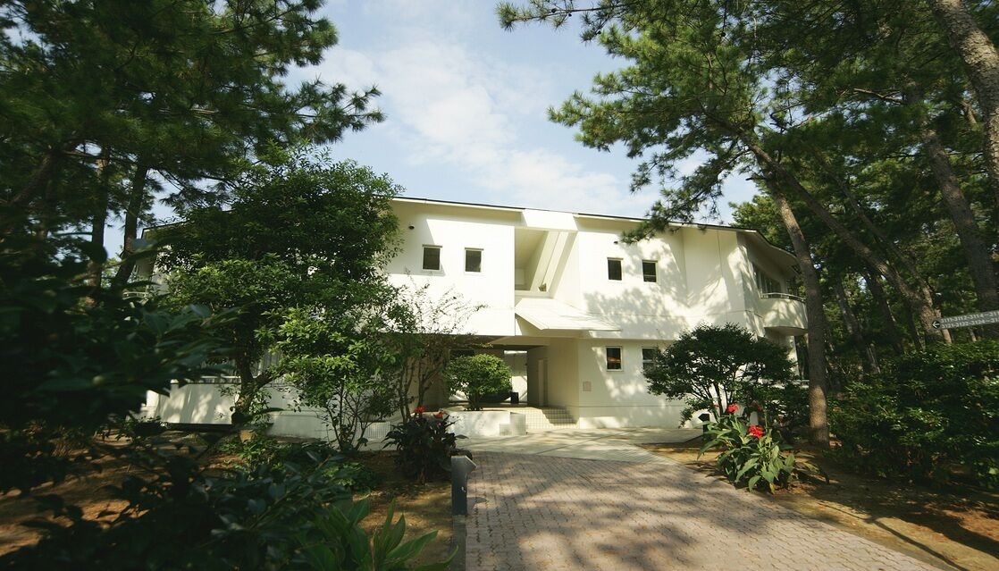 Un bâtiment à deux étages, doté d’un toit plat, entouré d’arbres et bordé d’une allée pavée.