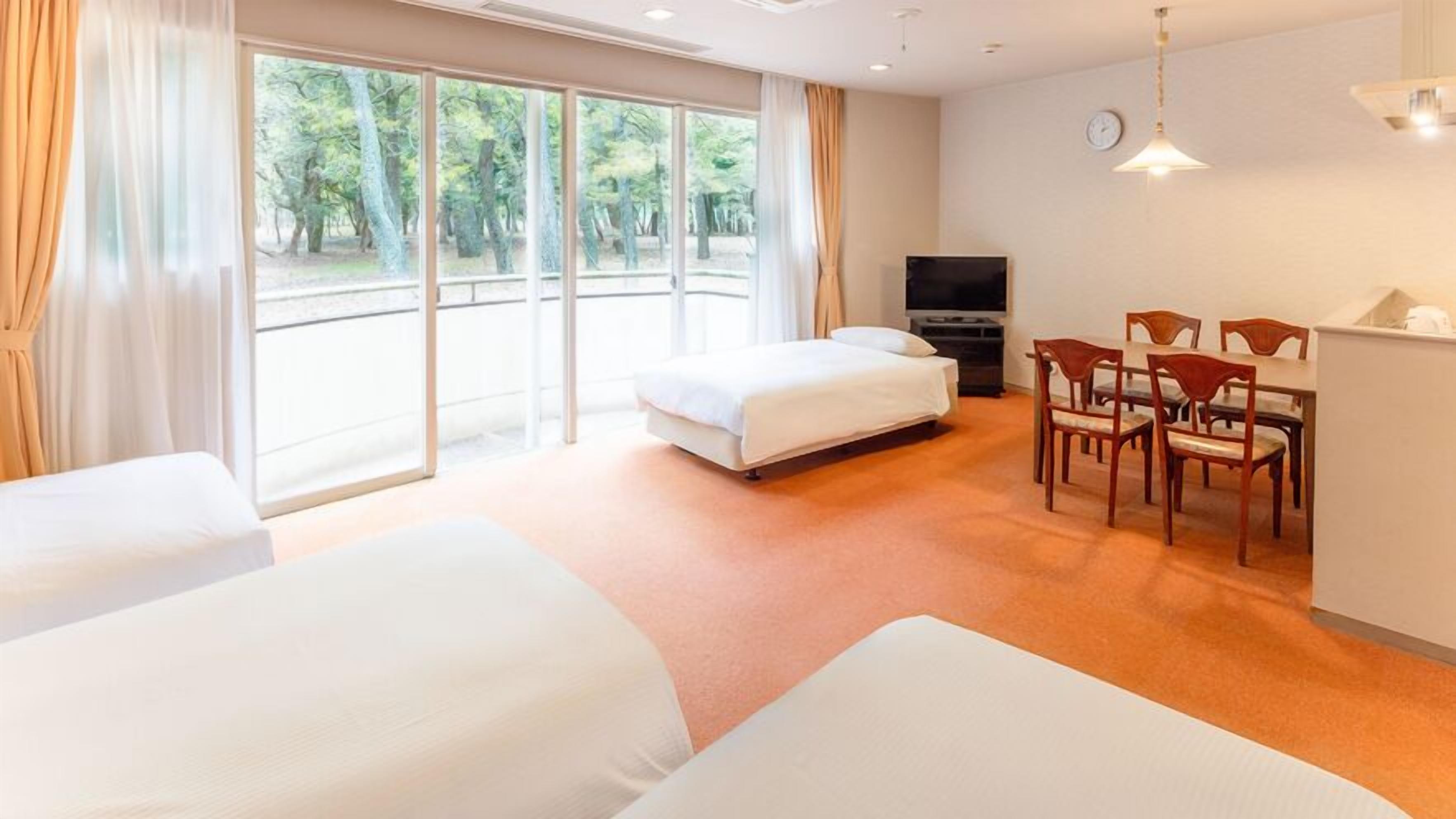 A hotel room with two beds, a dining area with chairs, a TV, and large windows with a view of trees.