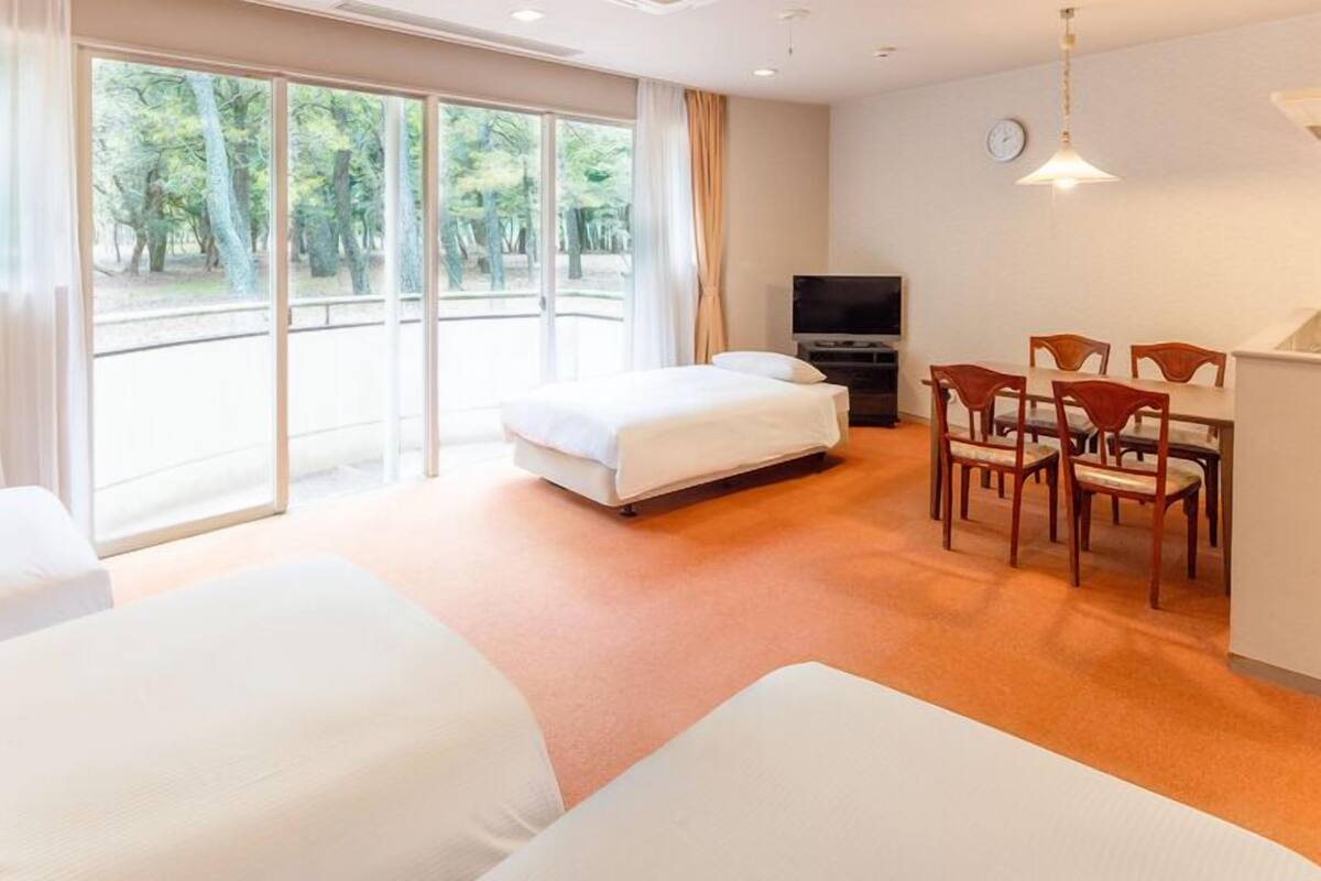 A hotel room with two beds, a dining area with chairs, a TV, and large windows with a view of trees.