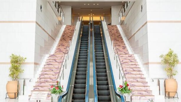 A modern hotel lobby with a central escalator flanked by stone steps, a reception desk, and potted plants. - Cottage Himuka (Miyazaki)