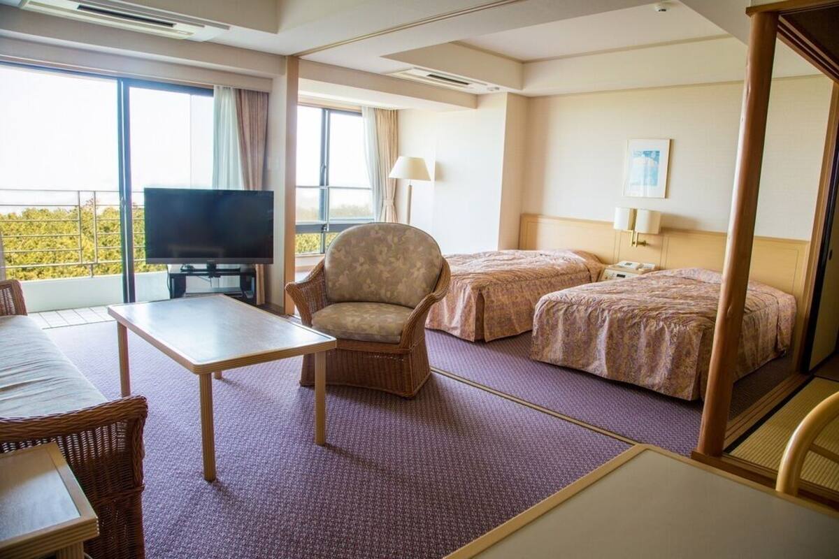 A hotel room with a TV, two beds, a chair, a table, and a balcony with a view.
