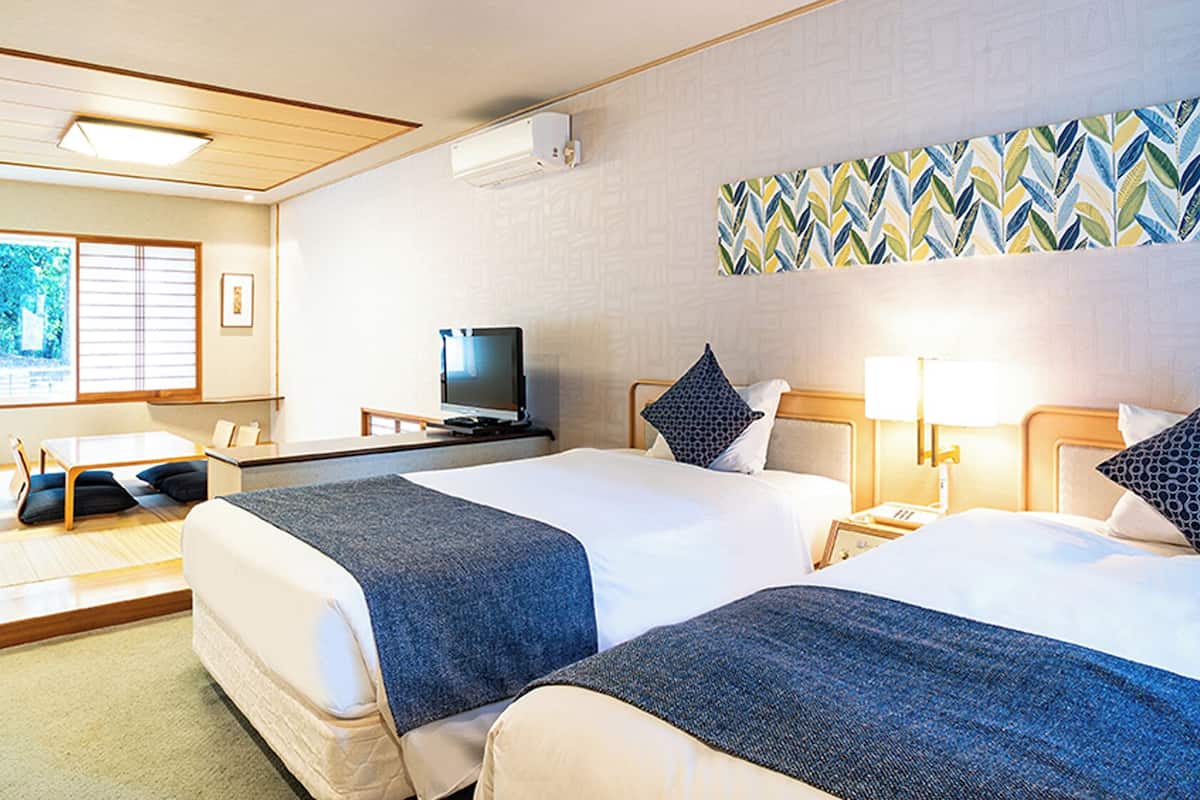 A hotel room with two beds, a desk, a television, and a large window with blinds.