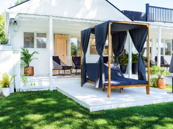 Un área de estar al aire libre con techo, con una cama con dosel, rodeada de plantas en macetas y sillas.