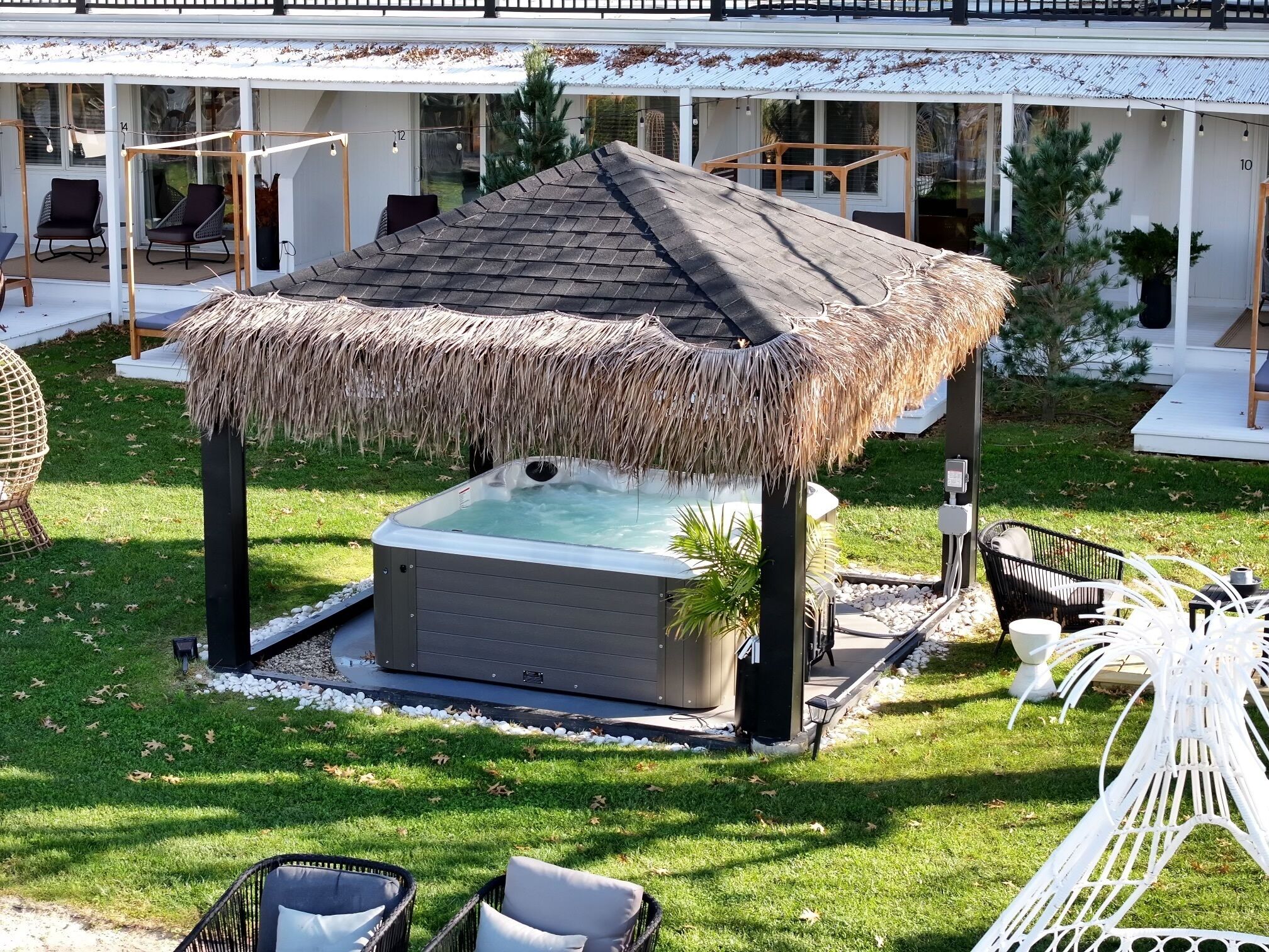 Un jacuzzi con techo de paja, rodeado de asientos exteriores y un edificio blanco al fondo.