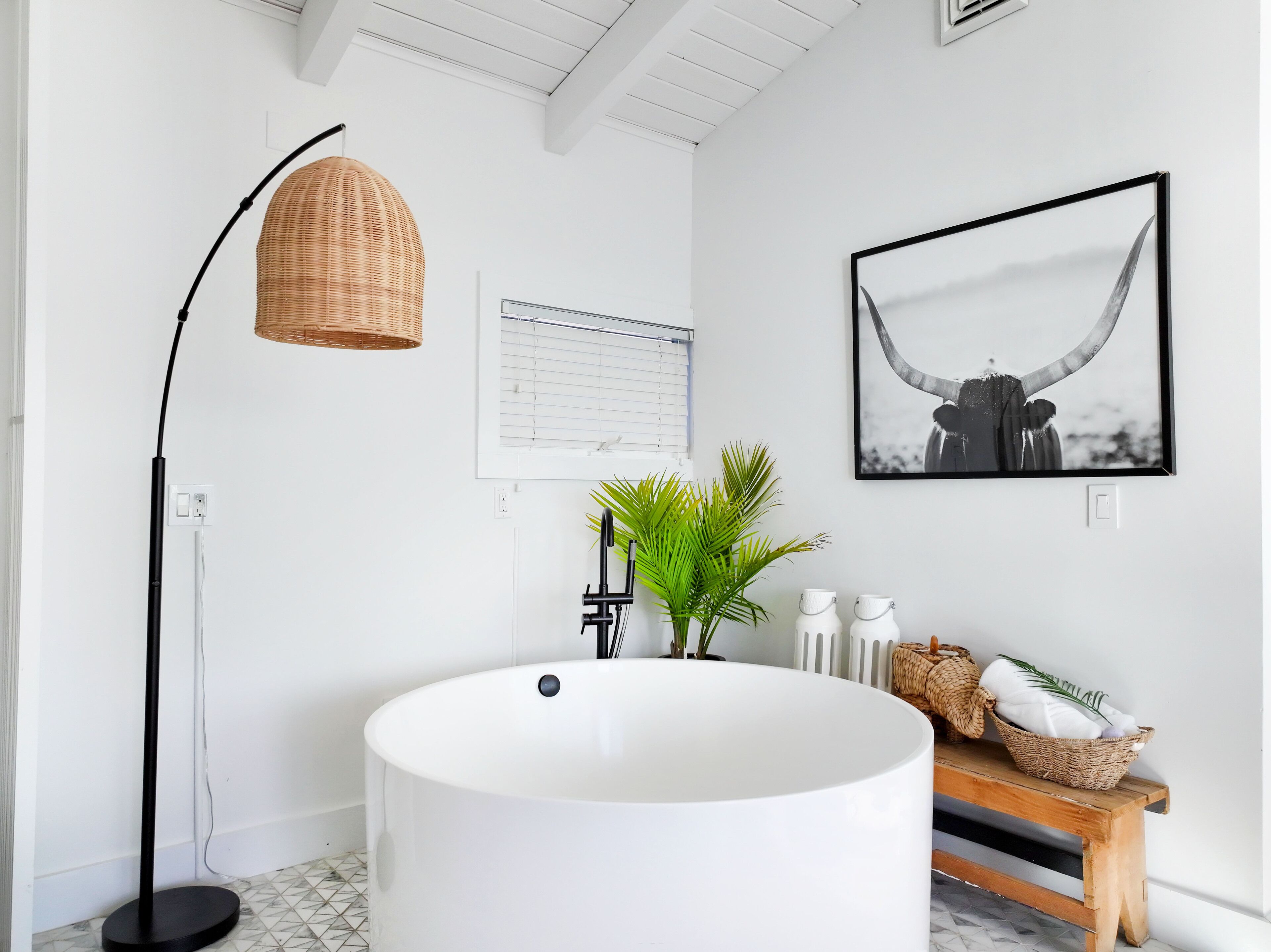 A modern bathroom with a freestanding floor lamp, a large tub, a potted plant, and a framed picture on the wall.
