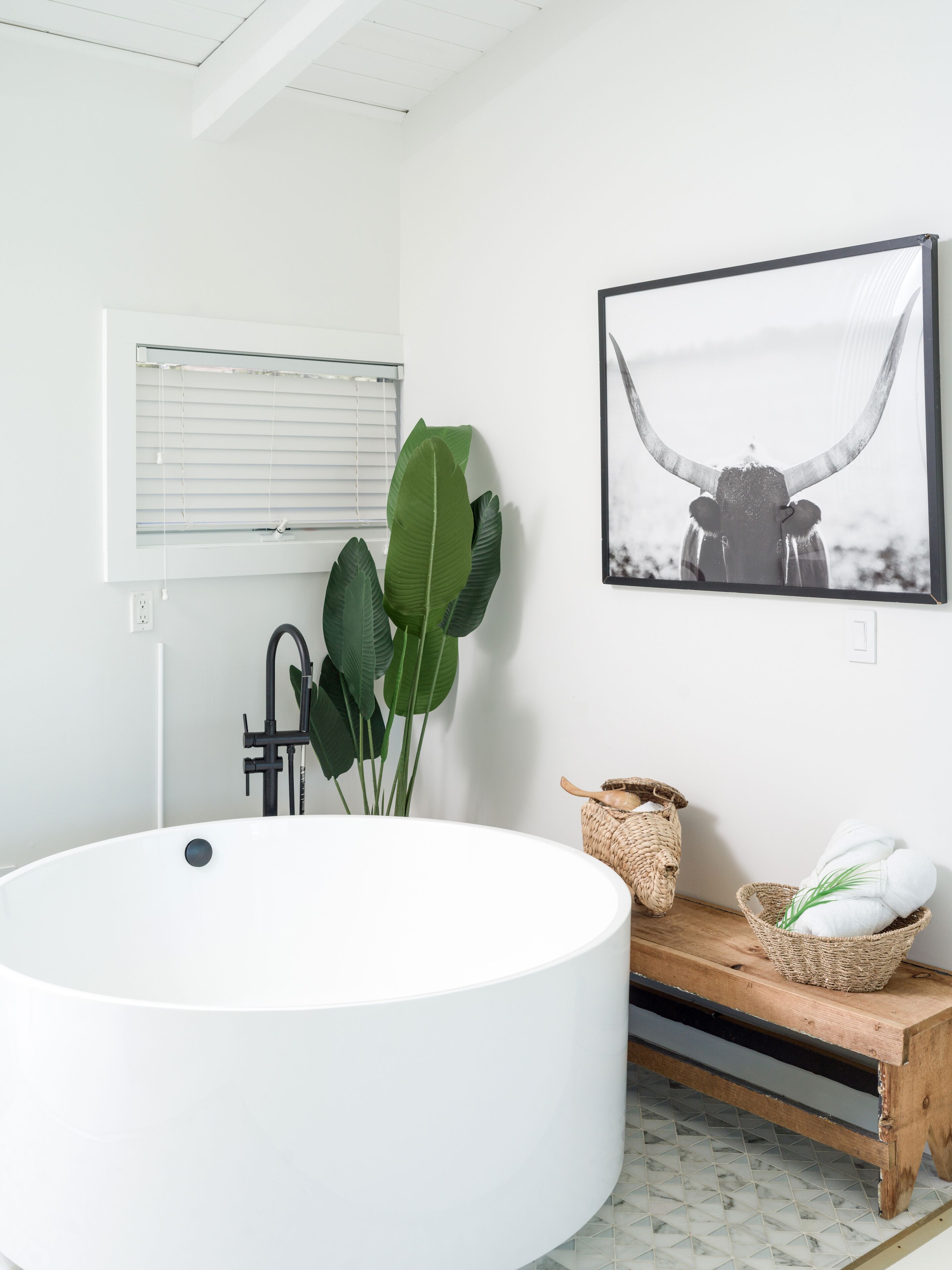 A modern bathroom with a large white bathtub, a black faucet, a large green plant, a woven basket, and a framed picture of a bull.
