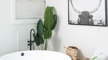 A modern bathroom with a large white bathtub, a black faucet, a large green plant, a woven basket, and a framed picture of a bull.