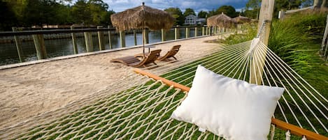 Un hamac avec un coussin, installé sur une pelouse près d’une étendue d’eau, des chaises longues en bois et des parasols en chaume.