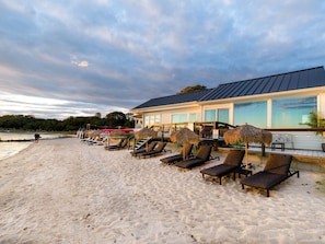 A beachside resort with sun loungers and thatched umbrellas, a covered seating area, and a building with large windows. - Aqua by American Beech - Adults Only (Riverhead)