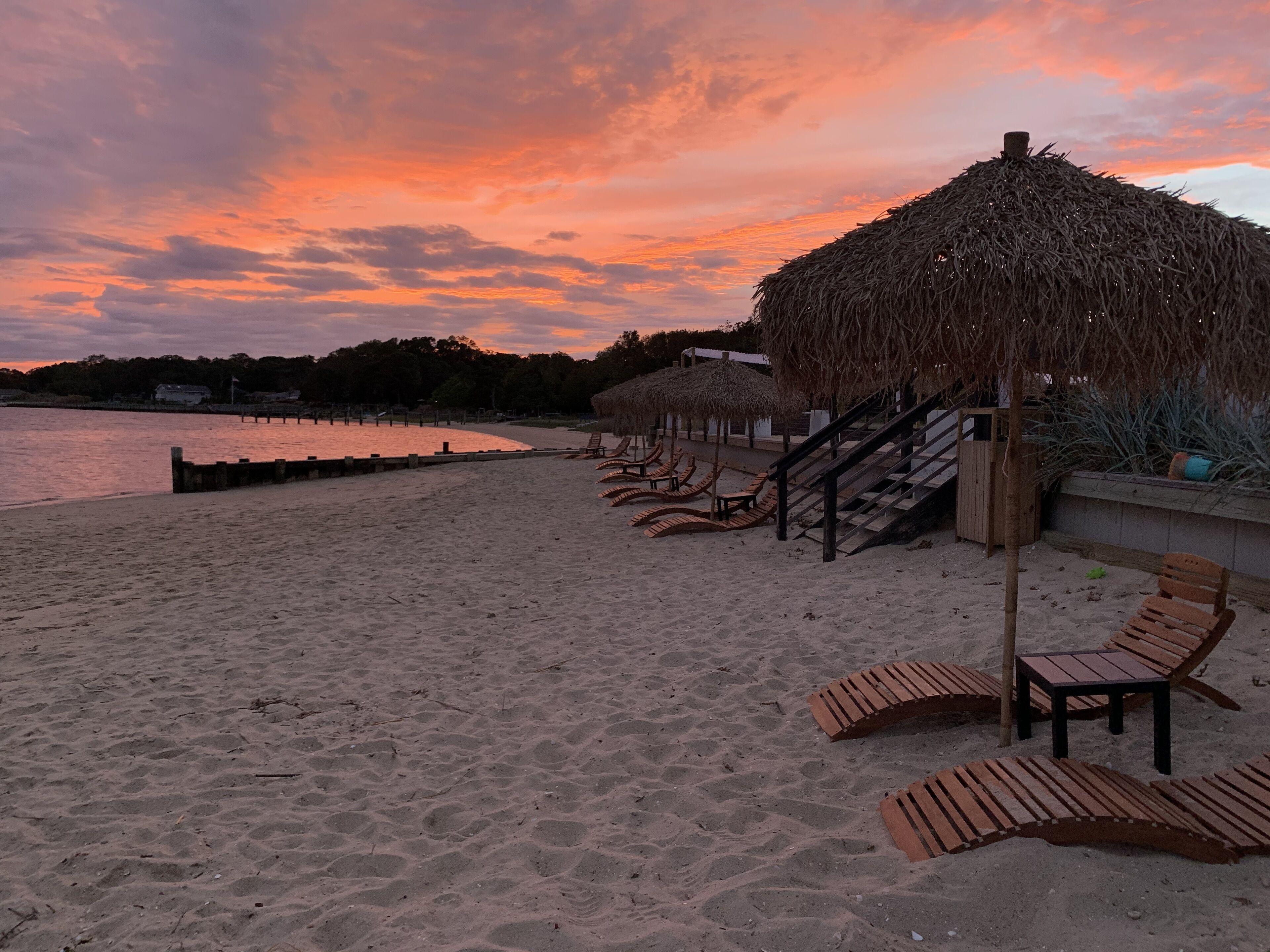 A beach with wooden lounge chairs and thatched umbrellas, a wooden staircase, and a view of the water.