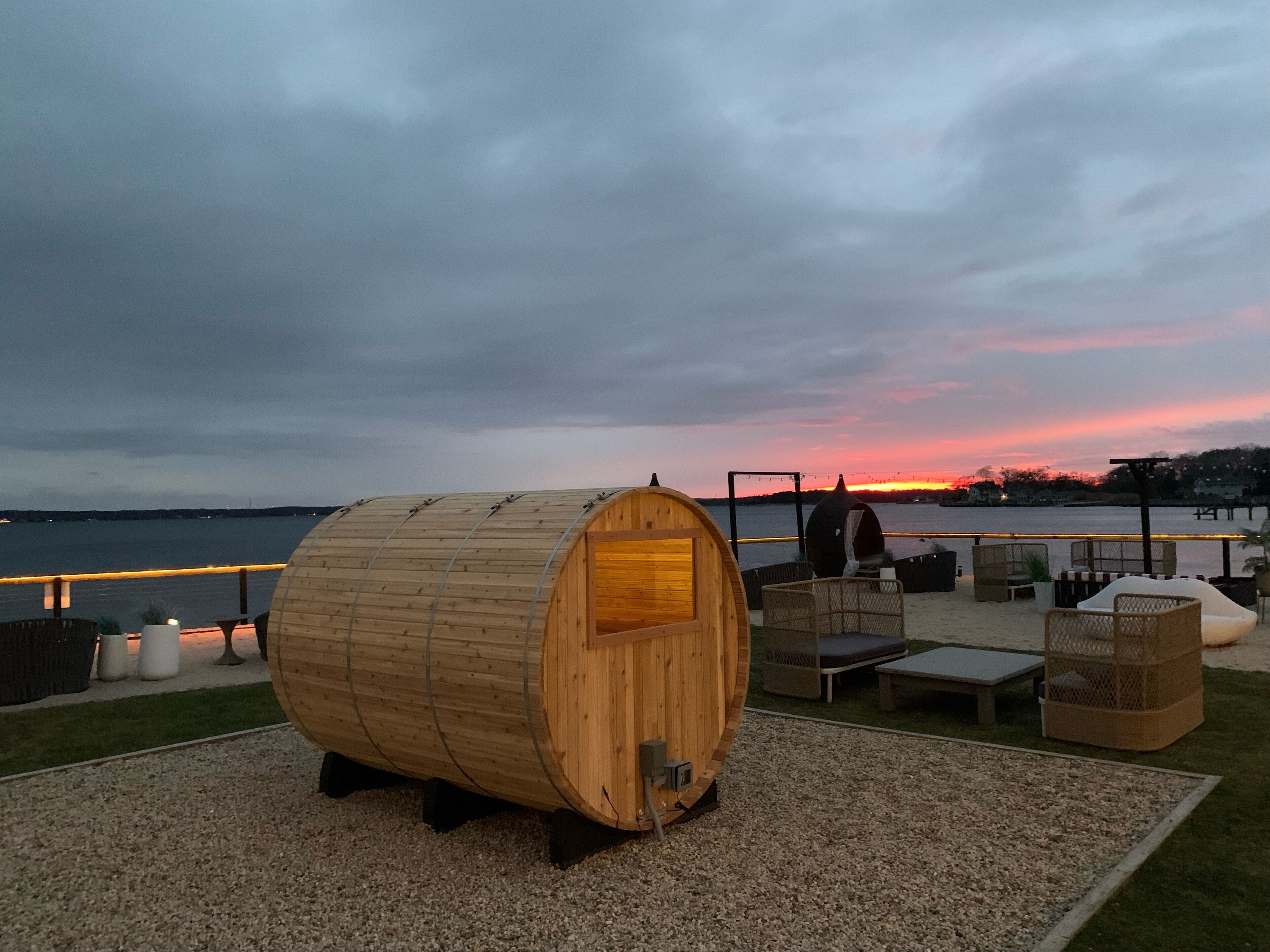 A wooden barrel structure with outdoor seating and a view of the sunset over water.
