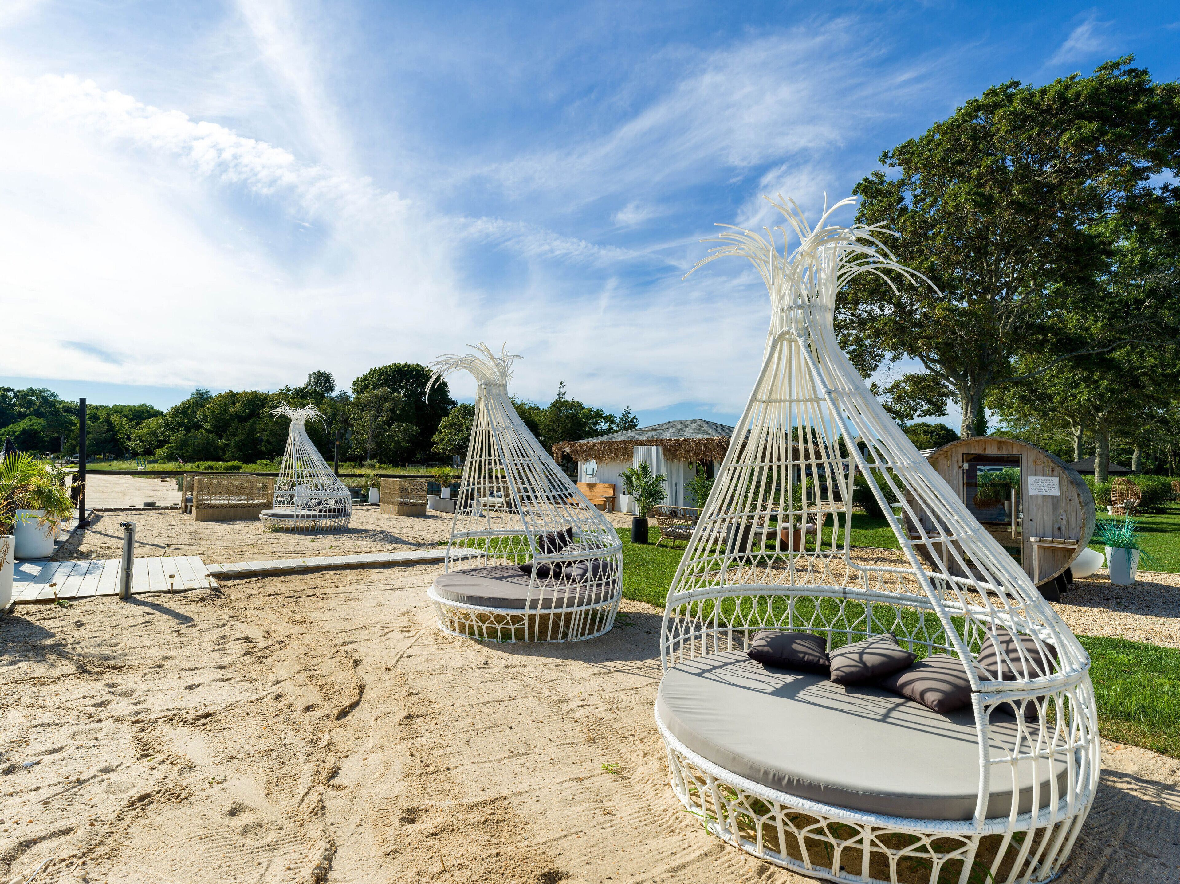 Outdoor seating areas with woven structures and cushions, surrounded by a sandy area and greenery.