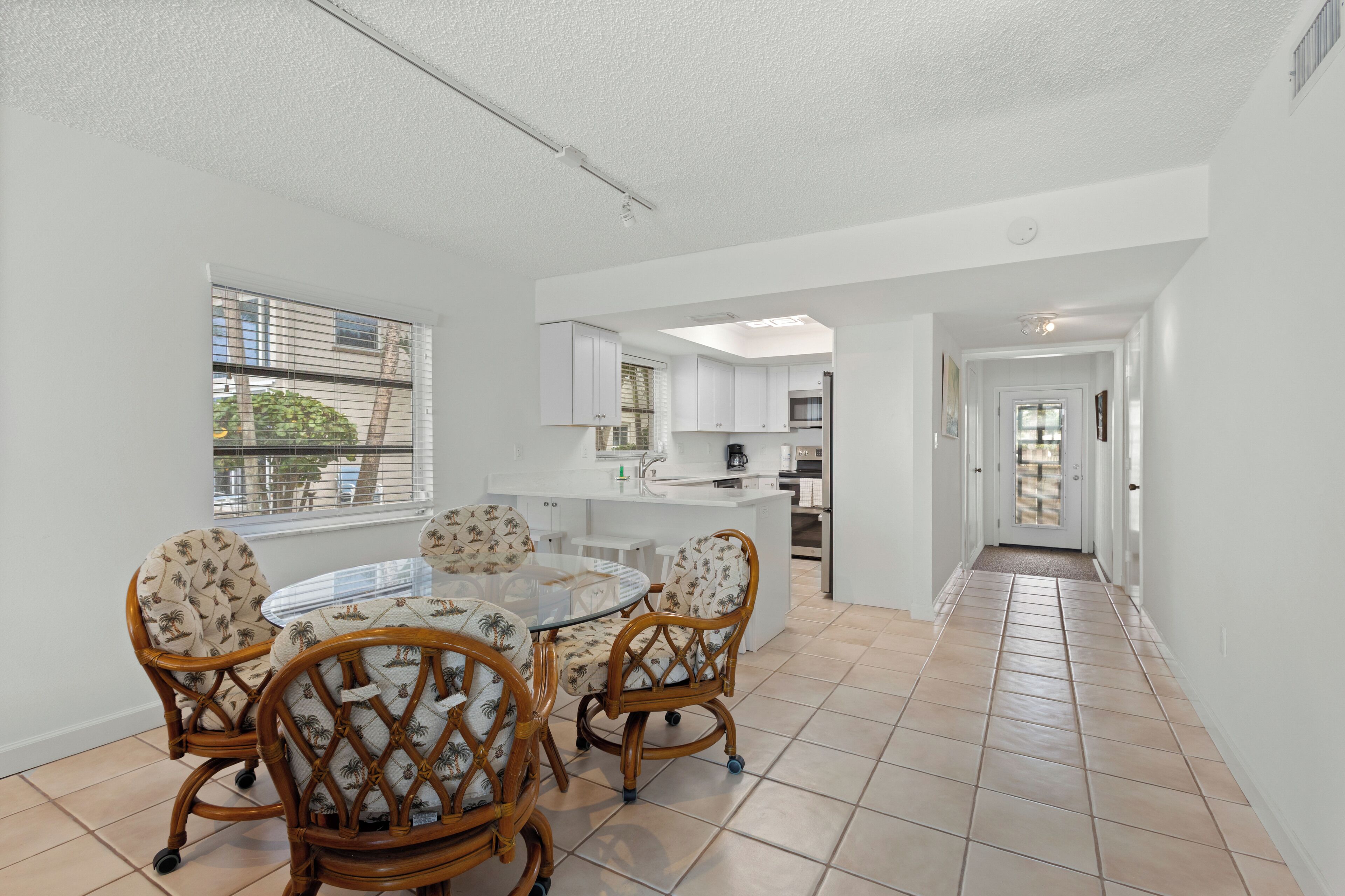 A spacious living area with a round glass table and wicker chairs, a kitchen with white cabinets, and a view through a window.