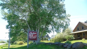 A sign for Cajun Cedar Log Cottages with a stone wall and a house in the background. - Cajun Cedar Log Cottages 2 Bdrm -  Unit 2 (Margaree Forks Cape Breton)