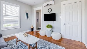 Ein Wohnzimmer mit Deckenventilator, einem an der Wand montierten Flachbildfernseher, Holzboden, einer weißen Tür und einem Fenster mit Jalousien.
