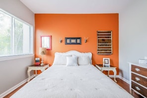 A bedroom with an orange wall, a white bed, a nightstand, a dresser, and a window with blinds.