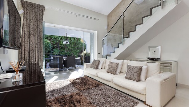 Un salon moderne avec un canapé blanc, un escalier en verre et une vue sur la verdure à l’extérieur.