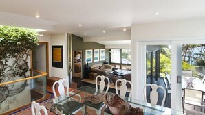 A modern interior with a glass dining table, white chairs, a sofa, and a large window offering a view of greenery. - BANNISTER COVE MOLLYMOOK - Art House - spa - (Mollymook Beach)