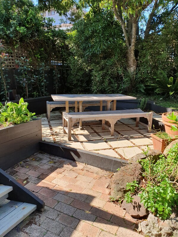 A wooden picnic table with benches in a garden area with brick flooring and surrounding greenery.