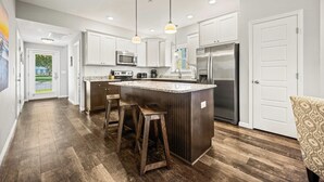 A modern kitchen with a central island, stainless steel appliances, and wooden bar stools. - Shore Beats Work, Modern 3 Bedroom Vacation Home w/ Community Pool & Clubhouse (Kill Devil Hills)