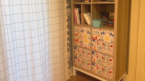 Huoneessa on puinen kaappi, jonka ovissa on värikkäitä laattoja, valkoinen verho ja puulattia.