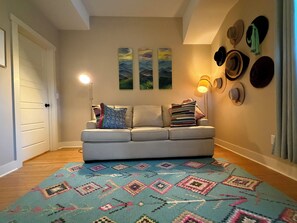 Ein Wohnzimmer mit einer Couch, einem gemusterten Teppich, an der Wand befestigten Hüten und gerahmten Kunstwerken.