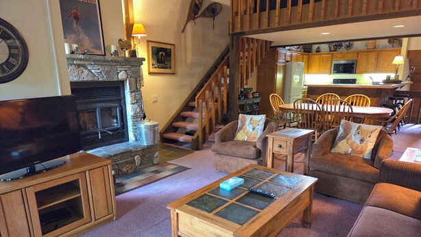 A cozy living room with a fireplace, comfortable seating, a television, and a staircase leading to a kitchen area. - Snowcreek Resort Vacation Rental 456 (Mammoth Lakes)