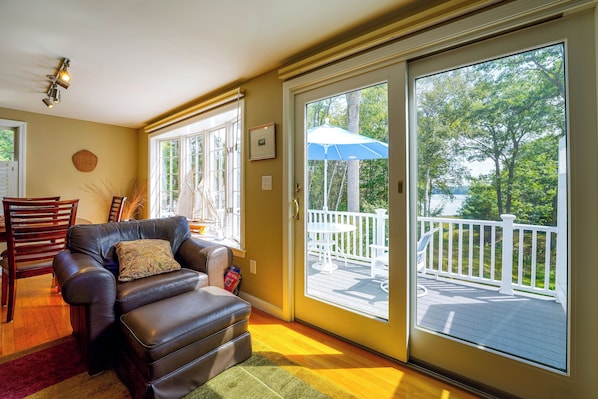 A living room with a leather sofa, a dining table, and a view of a deck and trees through large sliding glass doors.