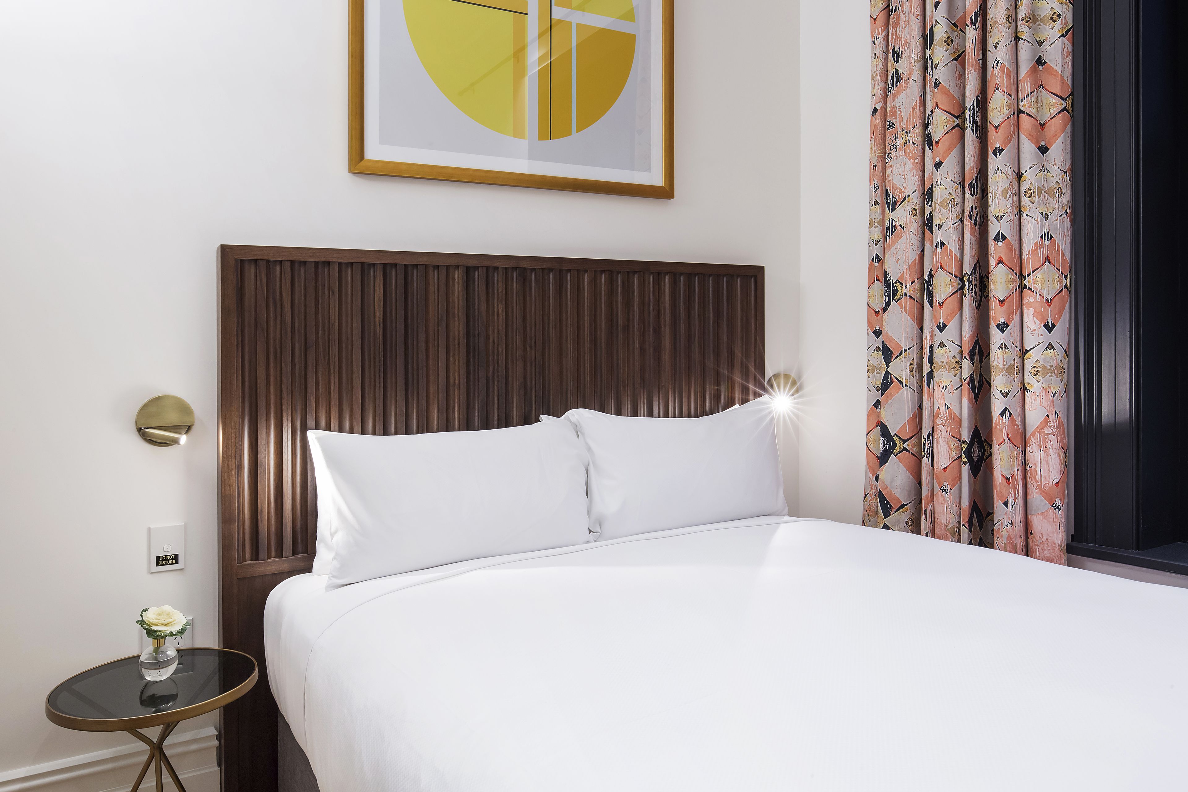 A neatly made bed with white linens, a wooden headboard, a small bedside table with a vase of flowers, and a wall-mounted light.