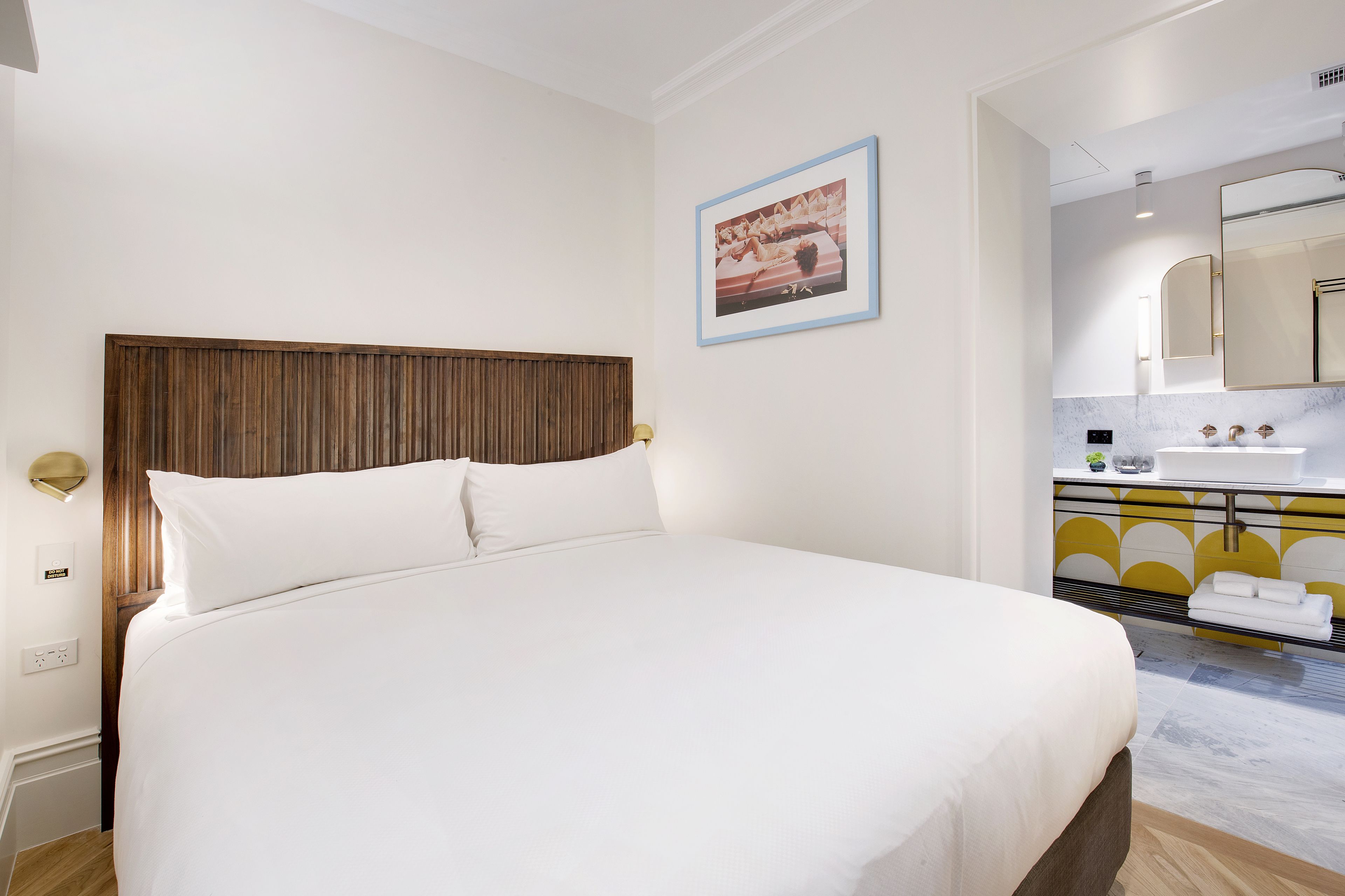 A hotel room with a large bed, a wooden headboard, a small bathroom with a mirror, and a picture on the wall.