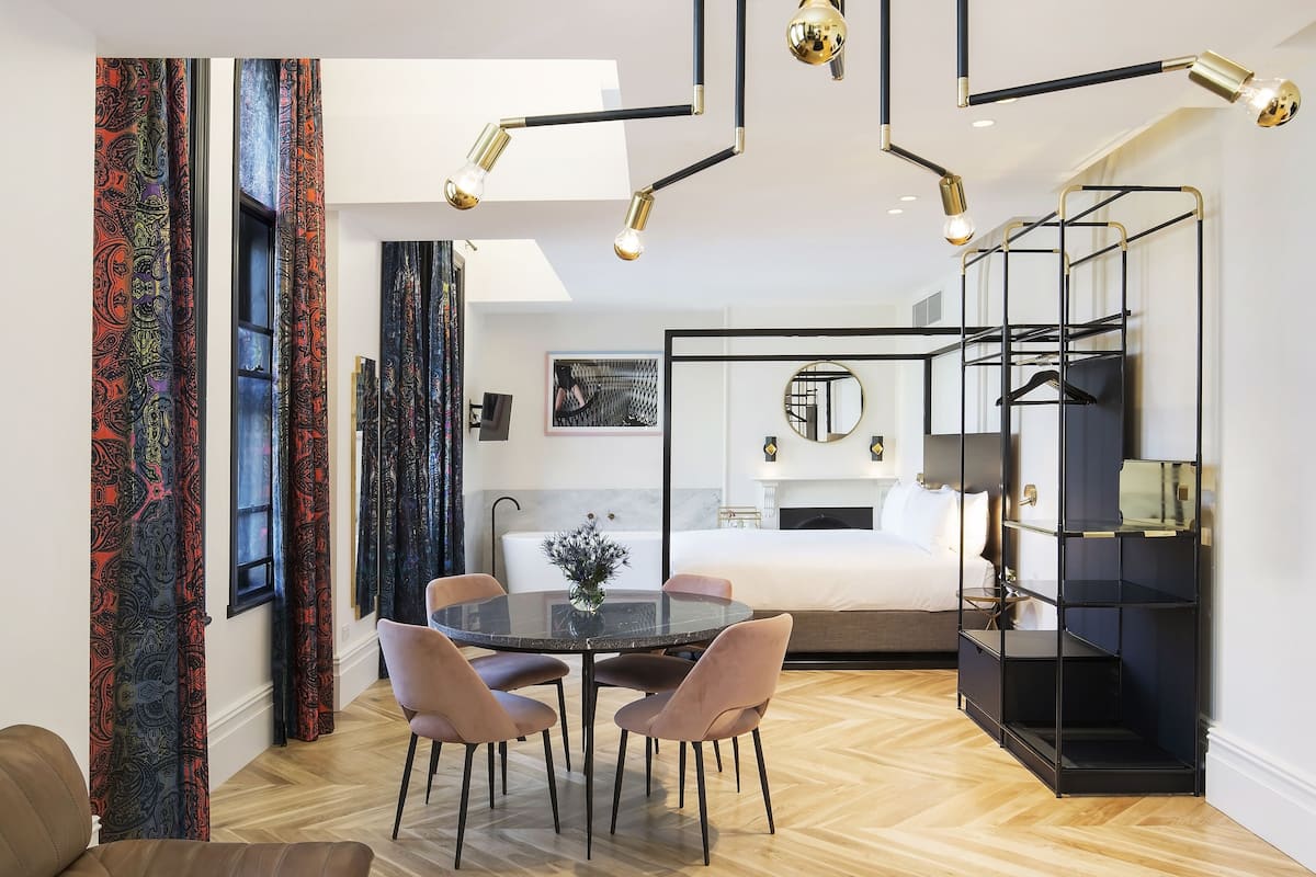 Una habitación de hotel moderna con una mesa de comedor de vidrio, dos sillas, un mueble negro para estantes y una cama con cabecera blanca.