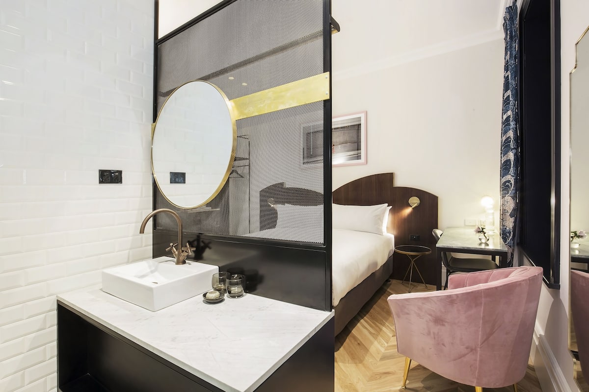 Una habitación de hotel moderna con una pared de azulejos blancos, un espejo redondo sobre un lavabo blanco, una cama con cabecero de madera, una mesita con una lámpara y una silla rosa.