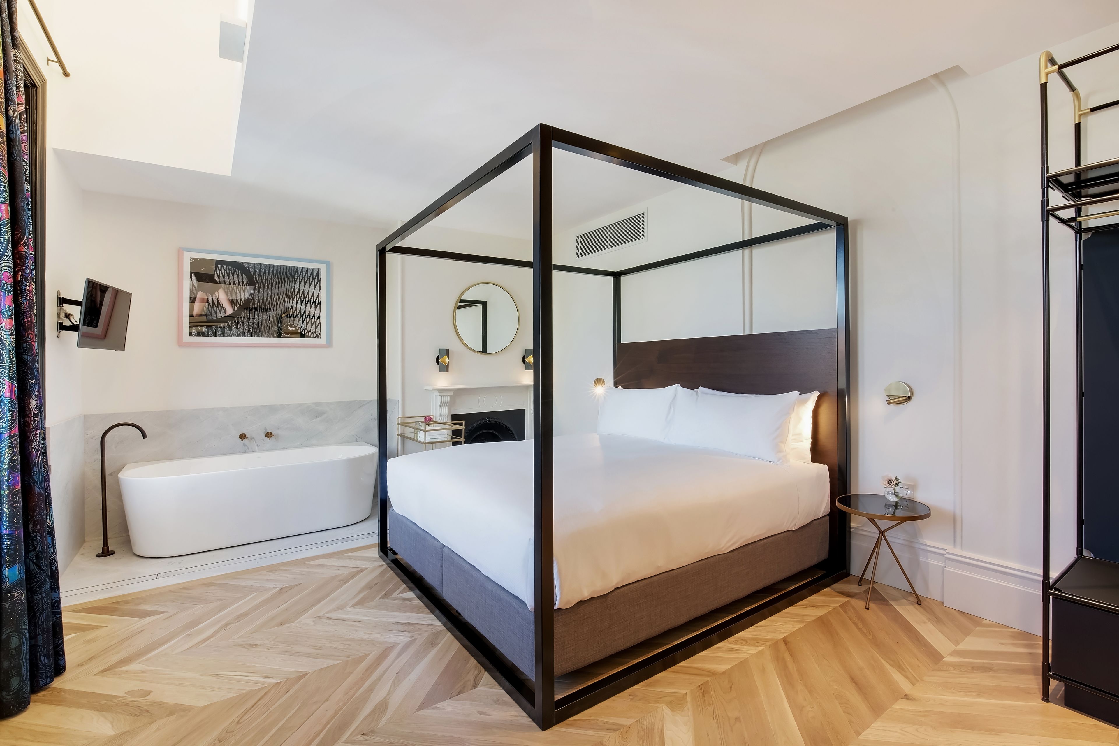 A modern bedroom with a glass four-poster bed, a separate bathtub, and a small bedside table.
