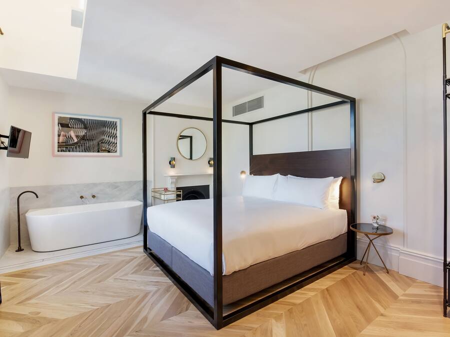 A modern bedroom with a glass four-poster bed, a separate bathtub, and a small bedside table.