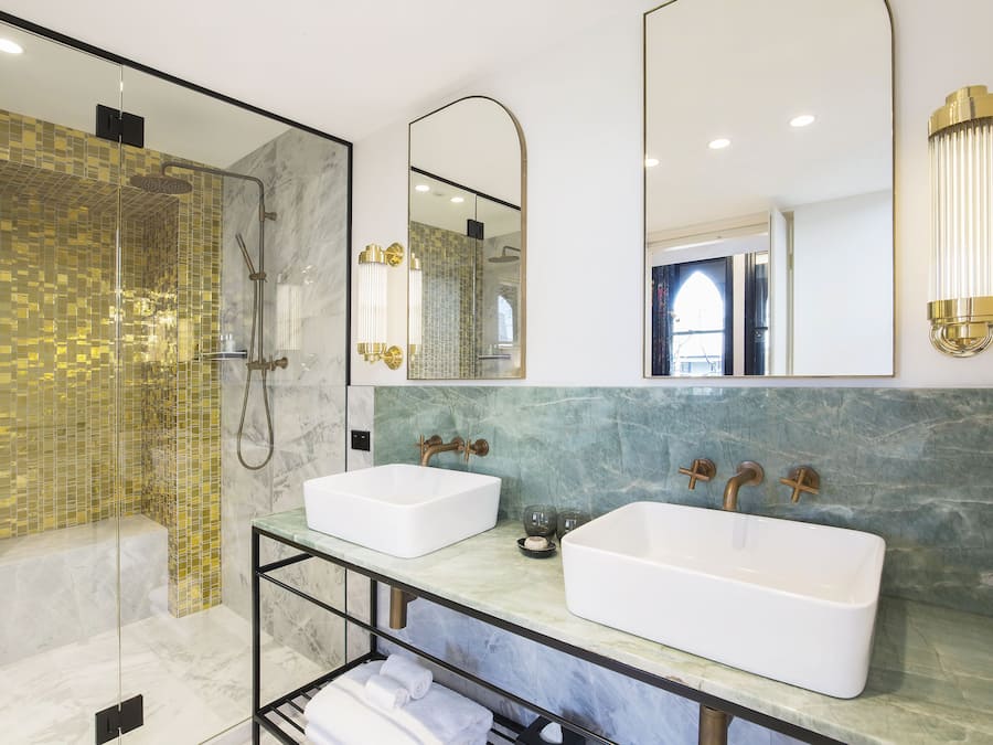 Un baño moderno con dos lavamanos, ducha con mampara de vidrio y salpicadero de mármol.