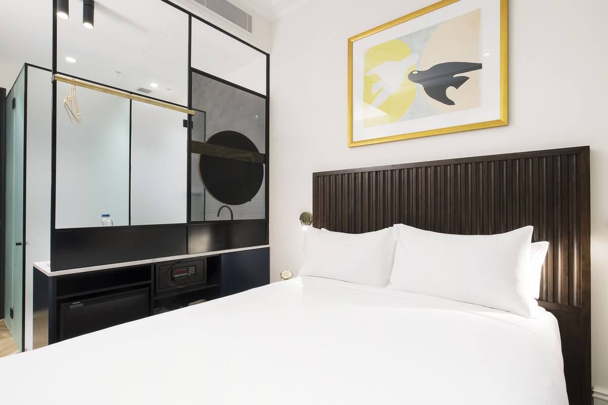 Una habitación de hotel con cama, un espejo grande, un cuadro abstracto enmarco y un televisor empotrado.
