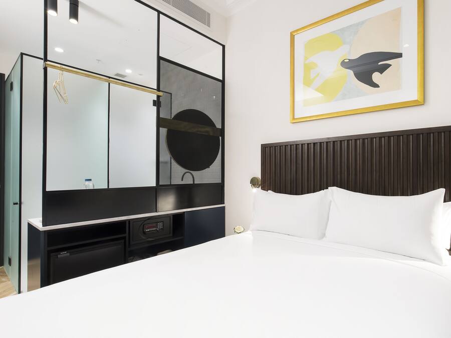 A hotel room with a bed, a large mirror, a framed abstract painting, and a built-in TV.