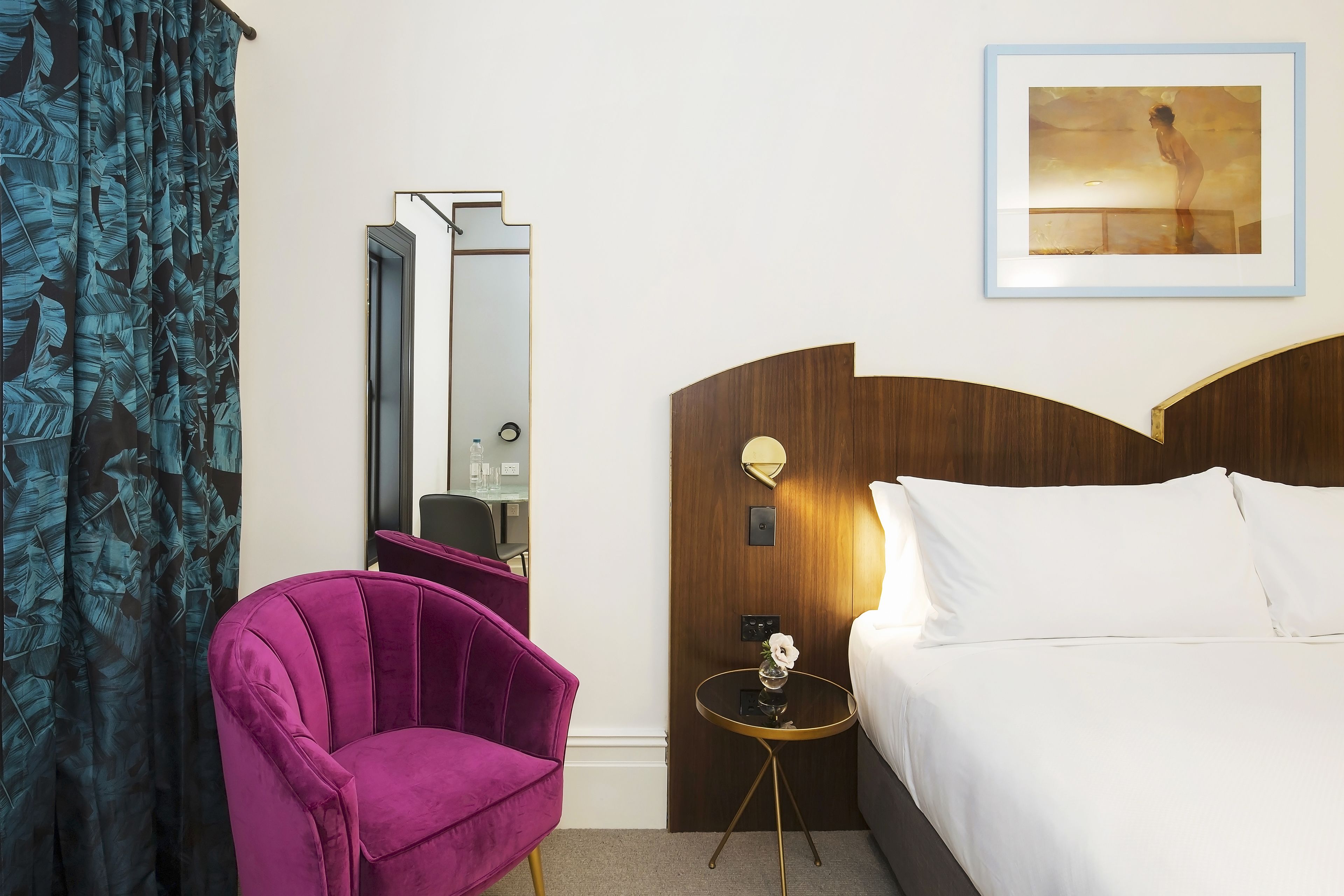 Habitación de hotel con cama, una silla morada, una mesita con un jarrón, un espejo y un cuadro en la pared.