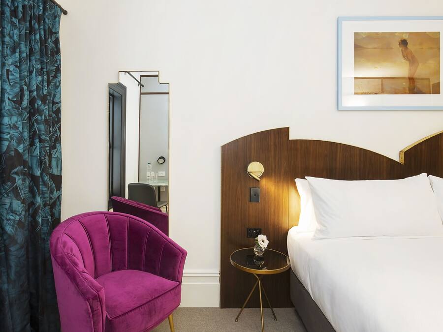 Habitación de hotel con cama, una silla morada, una mesita con un jarrón, un espejo y un cuadro en la pared.