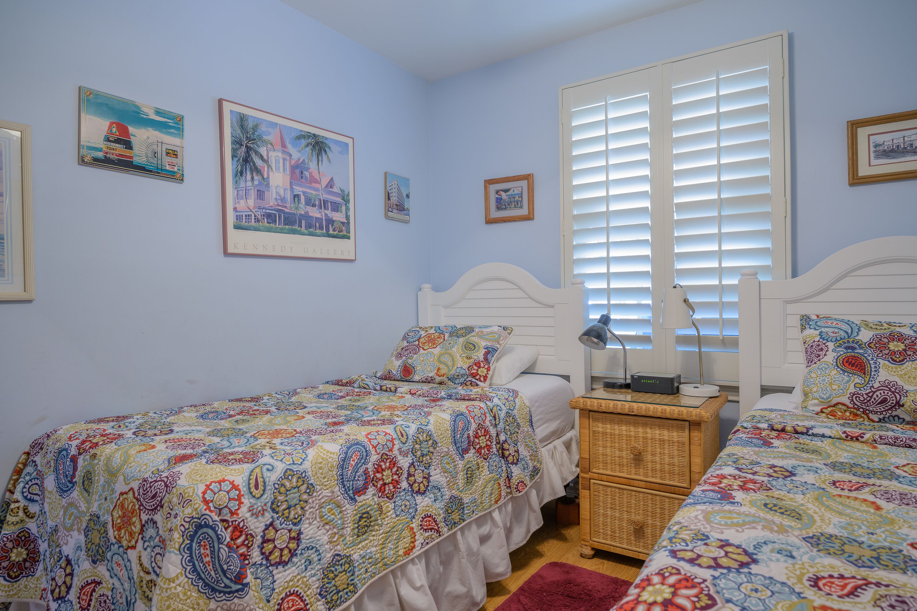A bedroom with two single beds, a nightstand, a wicker chair, and a window with blinds.