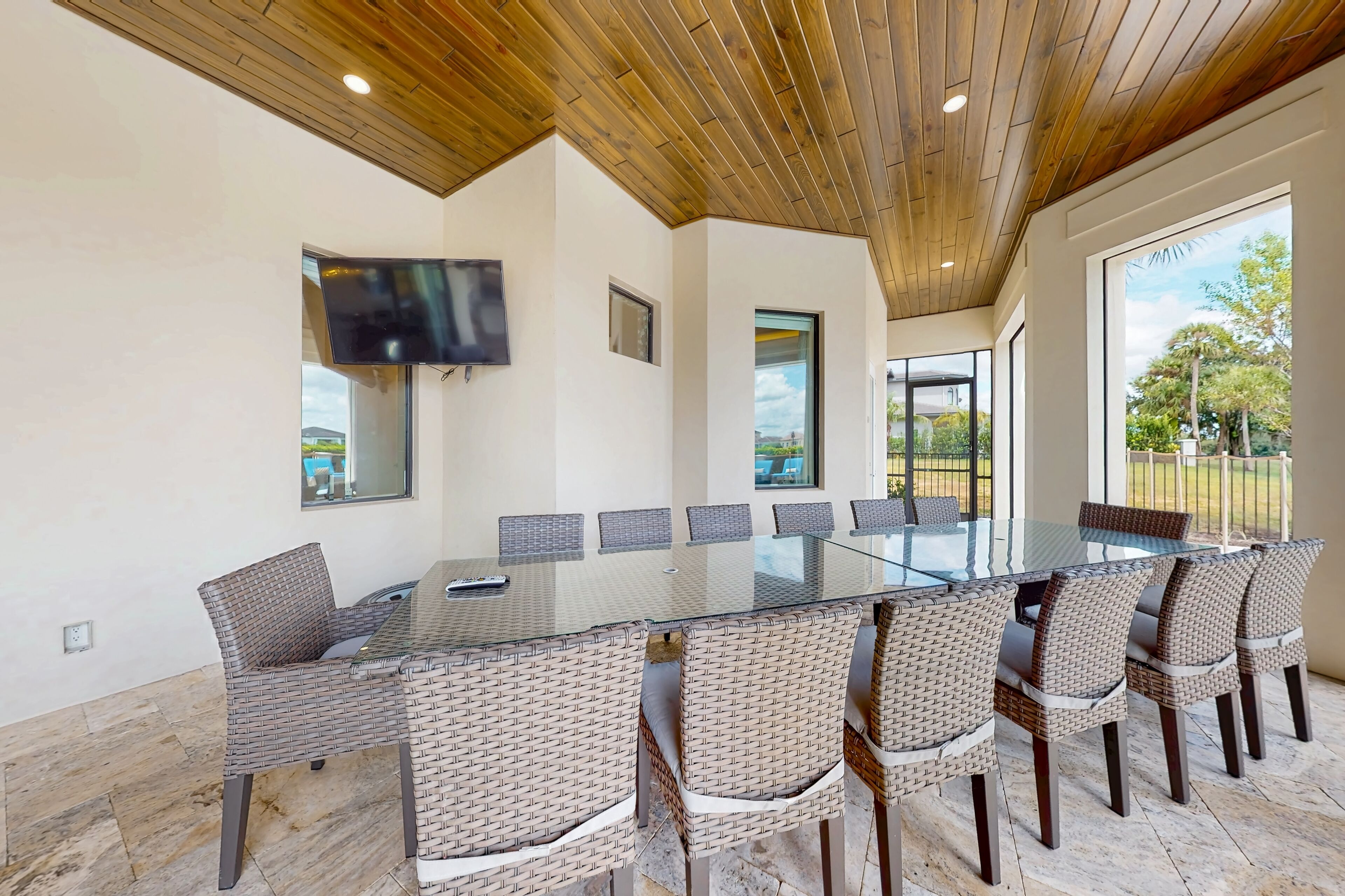 A spacious dining area with a glass table and wicker chairs, a mounted TV, and large windows offering a view of the outdoors.